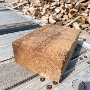 May include: A rectangular block of rough-hewn wood, showcasing a natural brown color and visible wood grain. The block sits on a corrugated metal surface, with a pile of chopped wood in the background. The wood block appears to be freshly cut.