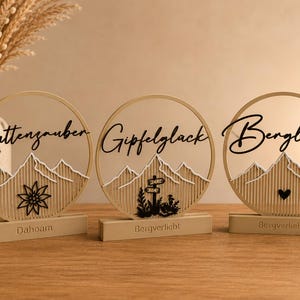 Puede incluir: Tres soportes decorativos circulares de madera con siluetas de montañas y texto. El izquierdo dice "Hüttenzauber" con una edelweiss, el central "Gipfelglück" con una señal, y el derecho "Bergliebe" con un corazón. Las bases dicen "Dahoam" o "Bergverliebt".
