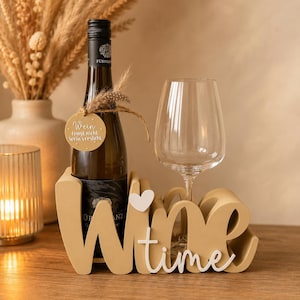 Puede incluir: Un soporte para botella de vino de color beige con forma de la palabra Wine, un corazón blanco y la palabra time en letra cursiva blanca. Incluye una botella de vino con una etiqueta redonda beige que dice Wein fragt nicht Wein versteht y una copa.