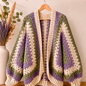 May include: A long-sleeved crochet cardigan featuring a geometric granny square pattern in alternating stripes of cream, sage green, and lavender. The open-front garment has a wide ribbed collar and cuffs, constructed with a textured yarn stitch.