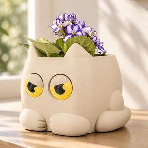 Op de afbeelding: Een beige katvormige plantenbak met grote, gele, opzij kijkende ogen en puntige oortjes. De plantenbak bevat een kleine plant met groene bladeren en paars-witte bloemen, waarbij de pootjes van de kat aan de onderkant rusten.