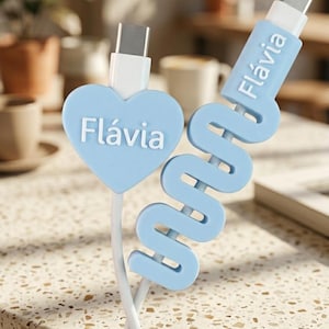 May include: A light blue charging cable accessory set featuring a heart-shaped cable protector and a wavy cable organiser, both personalised with the name Flávia in white lettering. The accessories are attached to white USB-C charging cables.