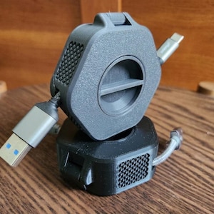 May include: Two dark gray hexagonal cable organizers with textured side panels and central circular winding mechanisms. Each device features a braided cable extending from the side, with one end showing a USB-A connector and the other a USB-C connector.