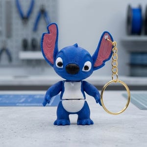 Peut inclure: Un porte-clés personnage bleu et blanc imprimé en 3D avec de grandes oreilles aux détails intérieurs roses, un nez noir et de grands yeux blancs. La figurine est fixée à un anneau métallique doré par une courte chaîne reliée à l'oreille gauche.