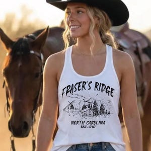 May include: A white tank top featuring a black graphic print of a mountain landscape with a cabin, pine trees, and the text "FRASER'S RIDGE NORTH CAROLINA EST. 1767". The garment has a scoop neckline and a racerback style, paired with blue denim jeans.