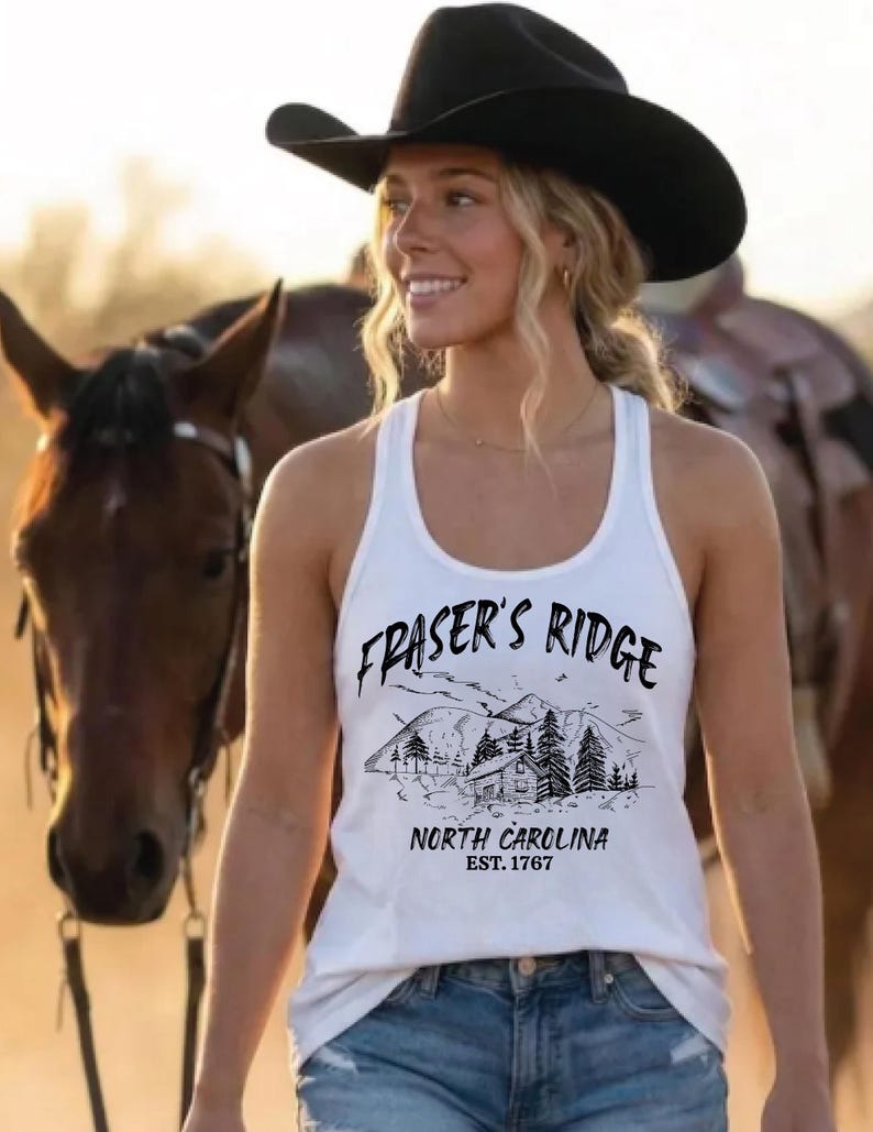 May include: A white tank top featuring a black graphic print of a mountain landscape with a cabin, pine trees, and the text "FRASER'S RIDGE NORTH CAROLINA EST. 1767". The garment has a scoop neckline and a racerback style, paired with blue denim jeans.