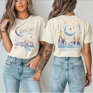May include: A cream-colored short-sleeve t-shirt featuring a graphic print of a city skyline under a crescent moon and stars. The front displays the text "CRESCENT City" while the back shows a larger version with the text "LUNATION".