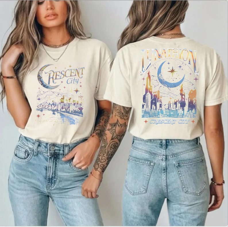 May include: A cream-colored short-sleeve t-shirt featuring a graphic print of a city skyline under a crescent moon and stars. The front displays the text "CRESCENT City" while the back shows a larger version with the text "LUNATION".