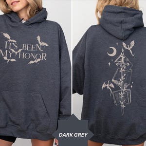 May include: A dark grey hooded sweatshirt featuring a front graphic with the text IT'S BEEN MY HONOR and small dragon silhouettes. The back displays a vertical design of a sword, open books, a crescent moon, and flying dragons.