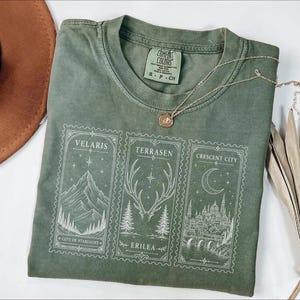 May include: An olive green crew neck t-shirt featuring three rectangular white graphic prints. The designs depict a mountain range labeled Velaris, a deer antler motif labeled Terrasen Erilea, and a city skyline labeled Crescent City.