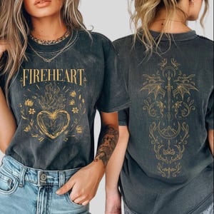 May include: Dark gray t-shirts with a gold design. The front of the shirt features the word "FIREHEART" above a heart engulfed in flames, surrounded by floral elements. The back of the shirt has a gold illustration of a sword, wings, and other decorative elements.