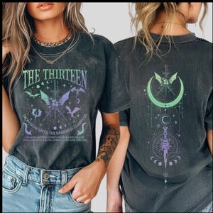 May include: Dark gray t-shirts with a graphic design. The front of the shirt features the text "THE THIRTEEN" and "FROM NOW UNTIL THE DARKNESS CLAIMS US" with bats, a sword, and other celestial elements. The back of the shirt has a crescent moon design.