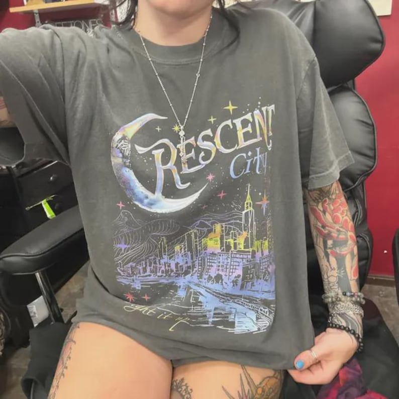 May include: A charcoal gray short-sleeve t-shirt featuring a graphic print of a crescent moon and a city skyline. The text Crescent City is printed in a stylized font above the skyline, accompanied by small star icons in shades of blue, purple, and yellow.
