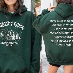May include: A dark green hooded sweatshirt featuring a white graphic on the front with a mountain cabin illustration and the text "FRASER'S RIDGE NORTH CAROLINA EST. 1767". The back displays the quote "YOU'RE BLOOD OF MY BLOOD, AND BONE OF MY BONE."