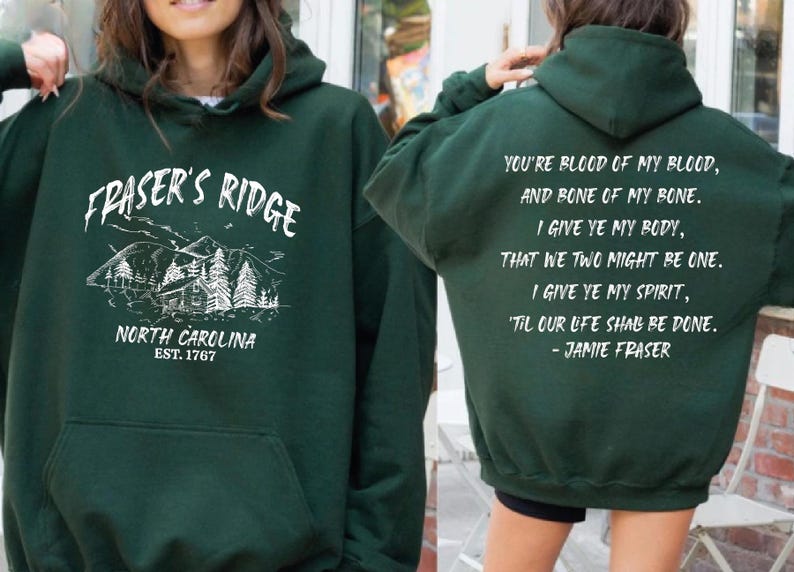 May include: A dark green hooded sweatshirt featuring a white graphic on the front with a mountain cabin illustration and the text "FRASER'S RIDGE NORTH CAROLINA EST. 1767". The back displays the quote "YOU'RE BLOOD OF MY BLOOD, AND BONE OF MY BONE."