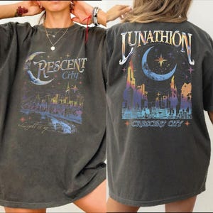 May include: A charcoal gray short-sleeve t-shirt featuring a graphic print of a city skyline under a crescent moon. The front displays the text Crescent City and Light it up. The back features the text LUNATHION and CRESCENT CITY.