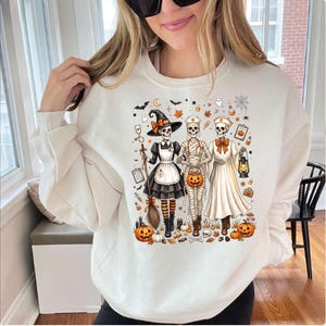 May include: A cream-colored crewneck sweatshirt featuring a graphic print of three skeletons dressed as a witch, a mummy, and a nurse. The design includes orange pumpkins, autumn leaves, bats, a spiderweb, and medical accessories like a stethoscope.