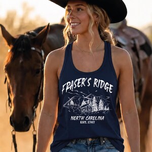 May include: A navy blue racerback tank top featuring a white graphic print of a mountain landscape with a cabin and pine trees. The text reads Fraser's Ridge, North Carolina, Est. 1767. The garment is paired with blue denim shorts and a black cowboy hat.