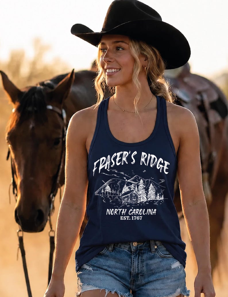 May include: A navy blue racerback tank top featuring a white graphic print of a mountain landscape with a cabin and pine trees. The text reads Fraser's Ridge, North Carolina, Est. 1767. The garment is paired with blue denim shorts and a black cowboy hat.