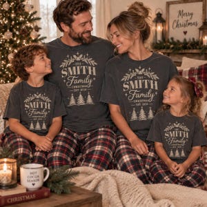 May include: A group of four people wearing matching charcoal gray t-shirts with a white graphic print that reads THE SMITH FAMILY CHRISTMAS EST. 2026, surrounded by pine branches and trees, paired with red, white, and black plaid pajama pants.