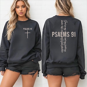 May include: A black long-sleeve sweatshirt featuring a white cross and the text Psalms 91 on the front chest. The back displays the text Psalms 91 in large letters with a vertical cross and the scripture God is my refuge and my fortress.