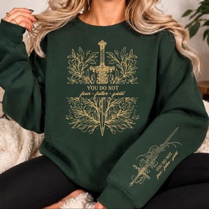 May include: A dark forest green crewneck sweatshirt featuring a gold-colored graphic of a sword surrounded by intricate leafy vines. The text "YOU DO NOT fear • falter • yield" is printed in the center and repeated on the left sleeve in gold.