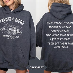 May include: A dark grey hooded sweatshirt featuring a white graphic on the front with the text Fraser's Ridge, North Carolina, Est. 1767 and a mountain cabin illustration. The back displays the white text You're blood of my blood, and bone of my bone.