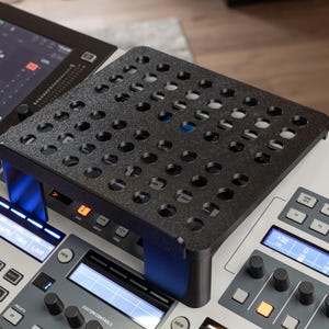 Może przedstawiać: Czarny, perforowany stojak znajduje się na konsoli mikserskiej audio Behringer. Stojak ma liczne okrągłe otwory i jest umieszczony nad przyciskami i ekranami konsoli. Konsola wyświetla różne ustawienia i elementy sterujące.