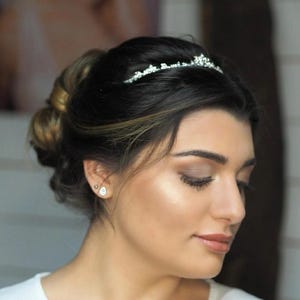 Puede incluir: Una persona con cabello oscuro recogido en un moño, adornado con una delicada tiara plateada con pequeños cristales transparentes. La persona viste una blusa blanca y pendientes en forma de lágrima. Un logo negro rectangular dice SPIEGELLIEBE VISAGISTIN.