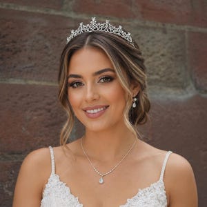 Puede incluir: Una tiara de tono plateado con intrincados adornos y cristales transparentes, combinada con un delicado collar de cadena plateada con un colgante en forma de lágrima y pendientes a juego, llevada con un vestido de encaje blanco.