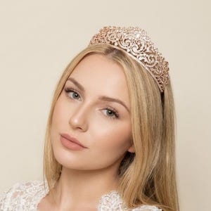 Puede incluir: Una tiara de oro rosa con un diseño intrincado y calado. La corona está adornada con detalles brillantes. La mujer de la imagen lleva un top de encaje blanco.