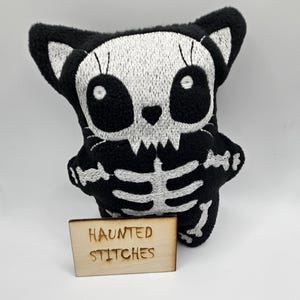 May include: A black and white plush cat toy with a skeleton design. The cat has a white embroidered skull face and rib cage on a black body. A small wooden sign with the text "HAUNTED STITCHES" is in front of the toy.