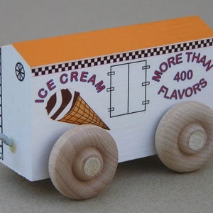 May include: A white wooden toy train car with an orange roof and natural wood wheels. The side features a graphic of an ice cream cone and the text "ICE CREAM MORE THAN 400 FLAVORS" with a black and white checkered border detail and a metal hook.