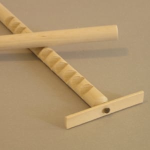 May include: Two wooden dowels, one with a carved pattern and a small wooden base, the other plain. The dowel with the pattern is resting on top of the plain dowel.