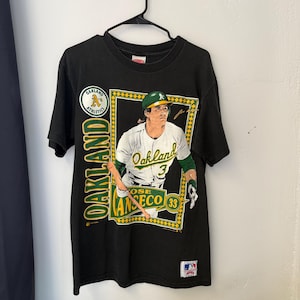 May include: Black t-shirt featuring a graphic of baseball player Jose Canseco in an Oakland Athletics uniform. The shirt includes the text "Oakland" and "Athletics" in green and yellow, with the player's name and number.
