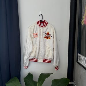 May include: Cream-colored satin jacket with red and white striped trim. The jacket features the text "Post 299" and a colorful emblem. The jacket is hanging on a white wall.