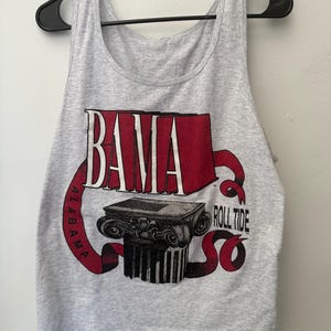 May include: A light gray sleeveless tank top featuring a graphic print with the word BAMA in red letters above a black and white illustration of a classical column. A red ribbon wraps around the design with the text ALABAMA and ROLL TIDE.