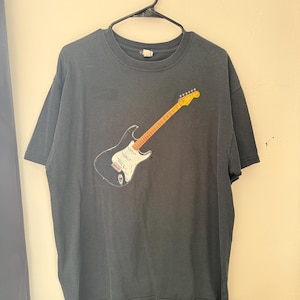 Pu&ograve; includere: T-shirt grigio scuro con una grafica di una chitarra elettrica. La chitarra ha un corpo nero, un battipenna bianco e un manico e paletta gialli. La maglietta &egrave; appesa a una gruccia nera contro un muro di colore chiaro.