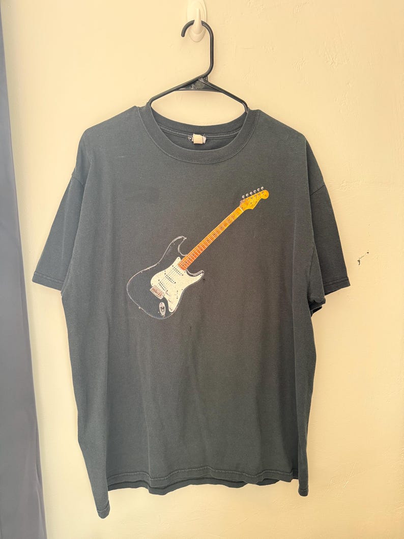 Pu&ograve; includere: T-shirt grigio scuro con una grafica di una chitarra elettrica. La chitarra ha un corpo nero, un battipenna bianco e un manico e paletta gialli. La maglietta &egrave; appesa a una gruccia nera contro un muro di colore chiaro.