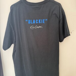 Pu&ograve; includere: T-shirt grigio scuro con la scritta "BLACKIE" in blu e la firma "Eric Clapton" in bianco. La maglietta &egrave; appesa a una gruccia nera. &Egrave; una maglietta a maniche corte e girocollo.