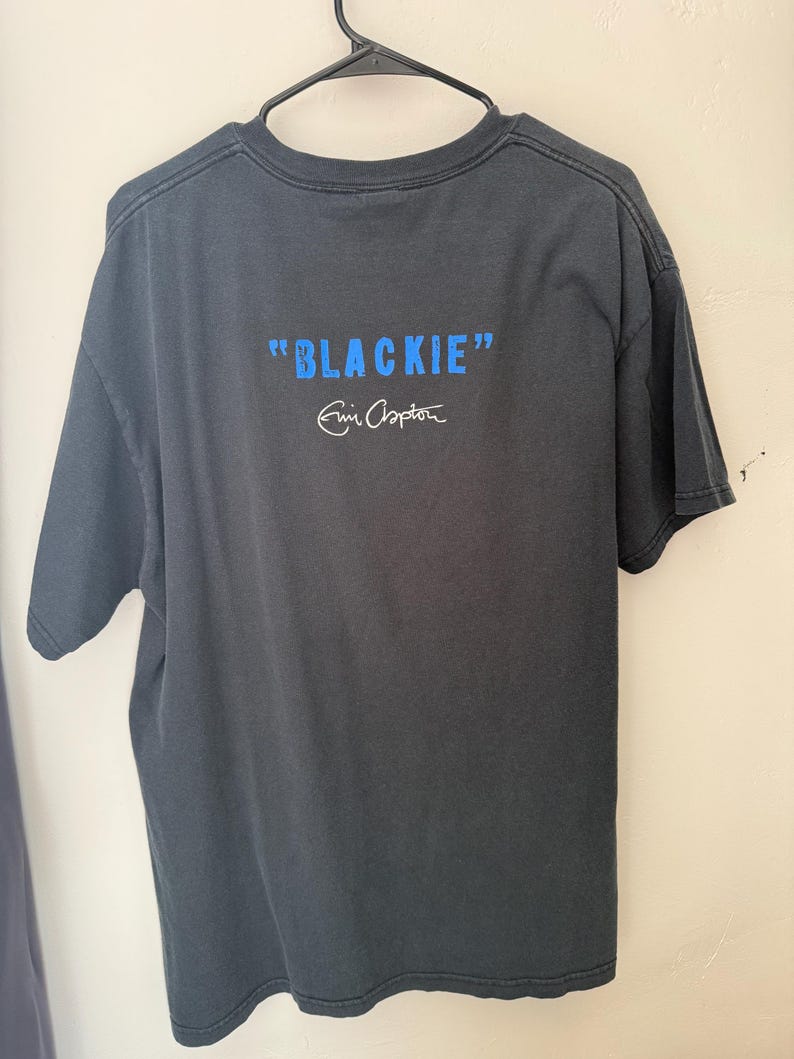 Pu&ograve; includere: T-shirt grigio scuro con la scritta "BLACKIE" in blu e la firma "Eric Clapton" in bianco. La maglietta &egrave; appesa a una gruccia nera. &Egrave; una maglietta a maniche corte e girocollo.