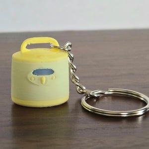 May include: A yellow and light green miniature rice cooker keychain. The keychain has a silver chain and ring. The rice cooker has a handle and a blue oval-shaped design on the front.