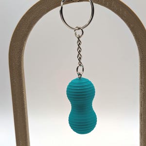 May include: A teal-colored, peanut-shaped keychain featuring a ribbed, layered texture. The object is attached to a silver-toned metal split ring by a short, twisted metal chain, creating a simple and functional accessory for keys or bags.
