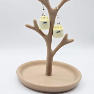 May include: A pair of dangle earrings featuring miniature pale yellow kettle charms with a small handle and a dark blue horizontal rectangular detail on the front. The charms are attached to silver-toned metal earring hooks with small jump rings.