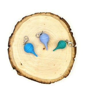May include: Three teardrop-shaped charms with a ribbed texture, each featuring a silver-toned lobster clasp. The charms are colored in shades of bright blue, light periwinkle, and teal, and they are arranged in a cluster on a light-colored surface.
