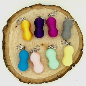 May include: A collection of eight peanut-shaped keychains in various solid colors including yellow, purple, magenta, gray, blue, light pink, mint green, and pale yellow. Each keychain features a silver-toned metal lobster clasp attachment.