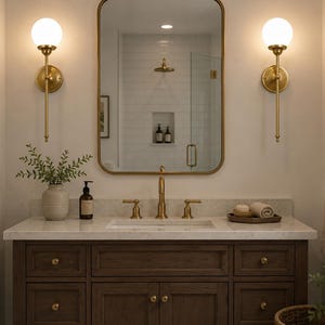 May include: A bathroom vanity featuring a dark wood cabinet with brass hardware, a white stone countertop, and an undermount sink. Above, a rectangular mirror with a brass frame is flanked by two wall-mounted brass globe light fixtures.