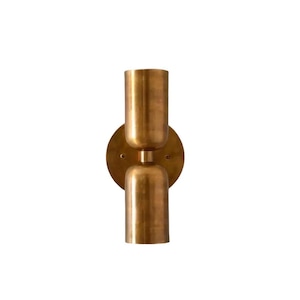 Peut inclure: Une applique murale avec une finition en laiton brossé. Le design présente une plaque arrière circulaire et deux douilles cylindriques ouvertes, disposées verticalement, l'une orientée vers le haut et l'autre vers le bas, reliées par une tige centrale.