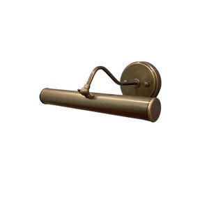 May include: A brass-toned picture light fixture. The fixture has a cylindrical light housing, a curved arm, and a circular wall mount. The finish is a warm, antique brass color, suitable for illuminating artwork or decor.