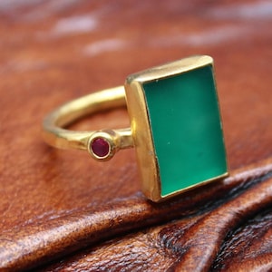 May include: A gold ring with a rectangular green stone and a small red stone accent. The ring is set on a brown leather surface, showcasing the jewelry's design and color contrast.